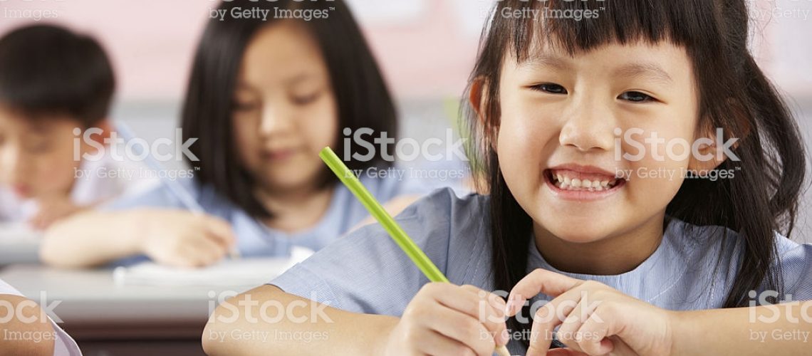 Young Female Student Working At Desks In Chinese School Classroom Smiling To Camera